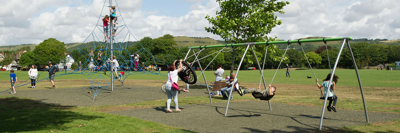 Seitenansicht eines großen Spielplatz-Schaukelgestells mit vielen Schaukelsitzoptionen, spielenden Kindern und Erwachsenen, die sie auf einem Spielplatz anschieben.